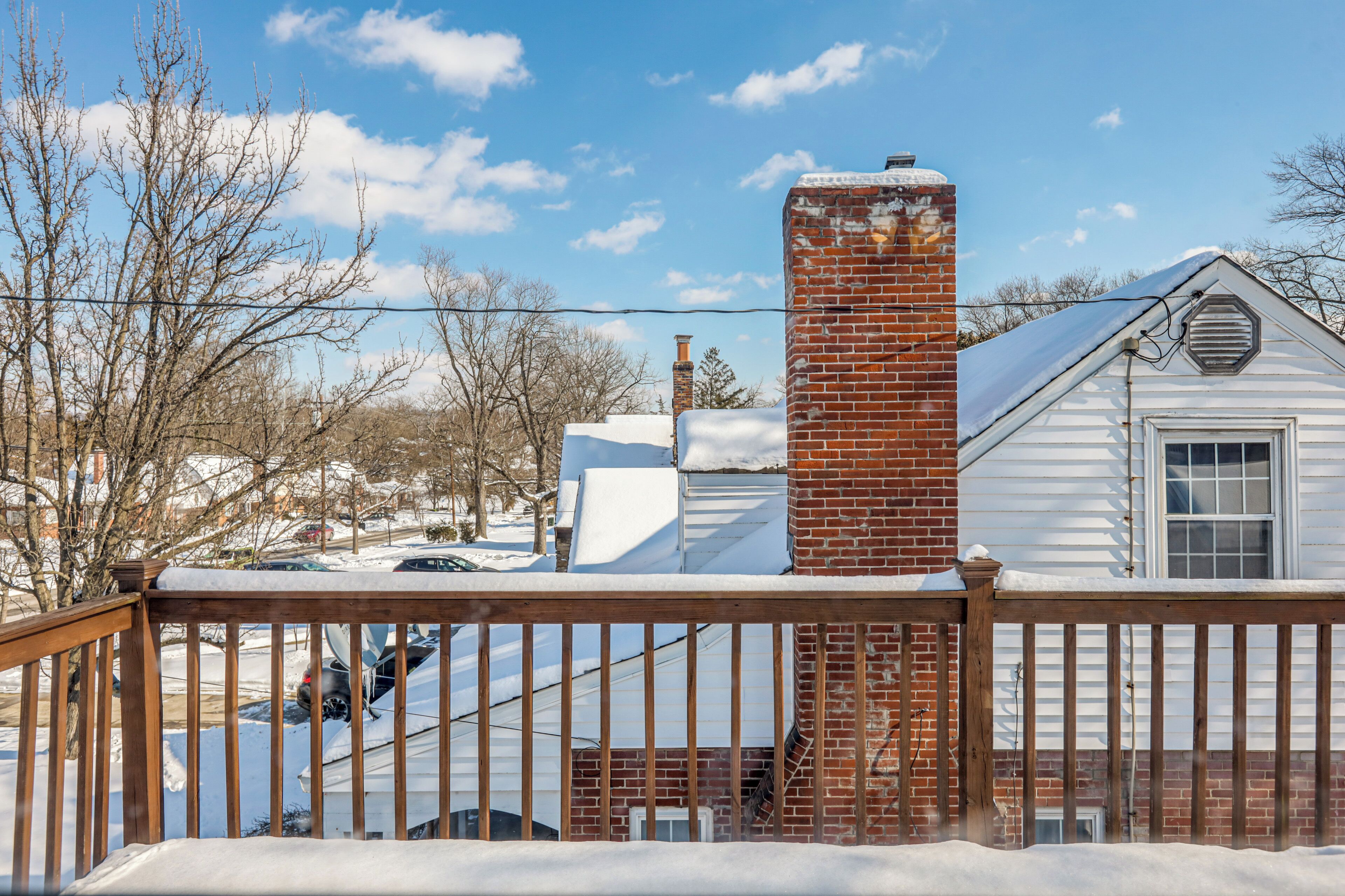 A wooden deck with a brick chimney, a white house with a window, and snow-covered surroundings.