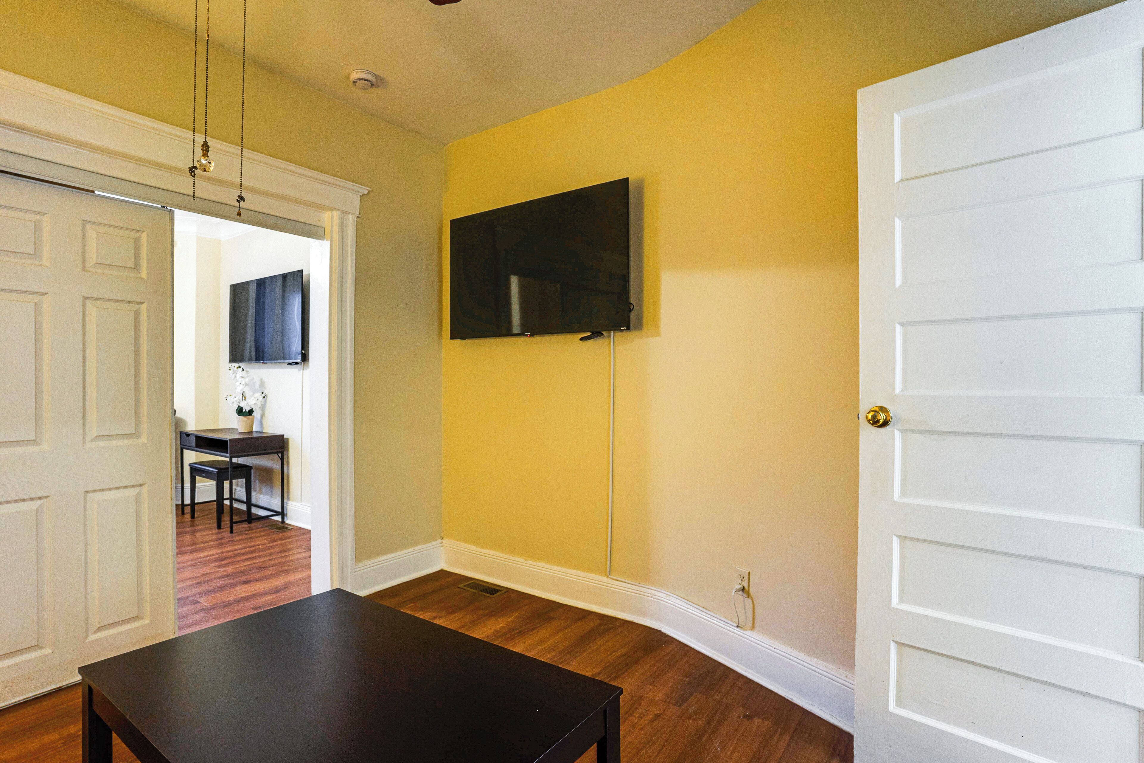 Una habitación con piso de madera, una mesa negra, una puerta blanca y un televisor instalado en la pared.