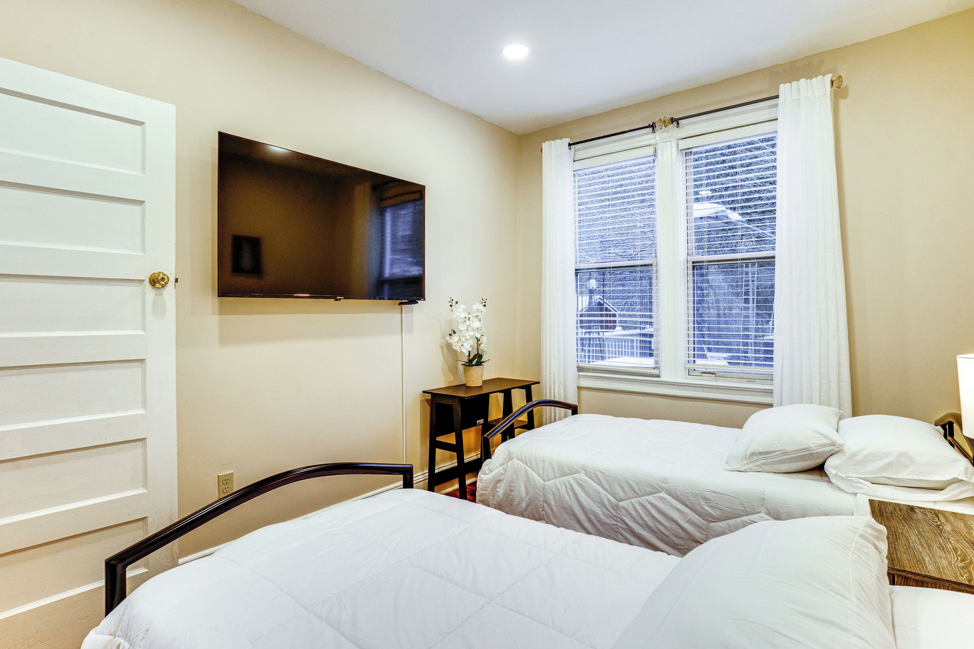 A hotel room with two beds, a TV mounted on the wall, a small table with a plant, and a window with blinds.