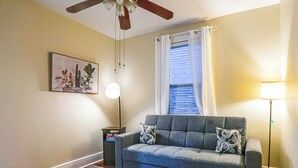 Un salon avec un canapé gris, deux coussins à motifs, un ventilateur de plafond, une lampe sur pied, une table d’appoint et une plante encadrée accrochée au mur.
