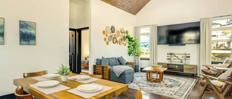 A spacious living room with a wooden dining table set for four, a comfortable sofa, a flat-screen TV, and a large window with curtains.