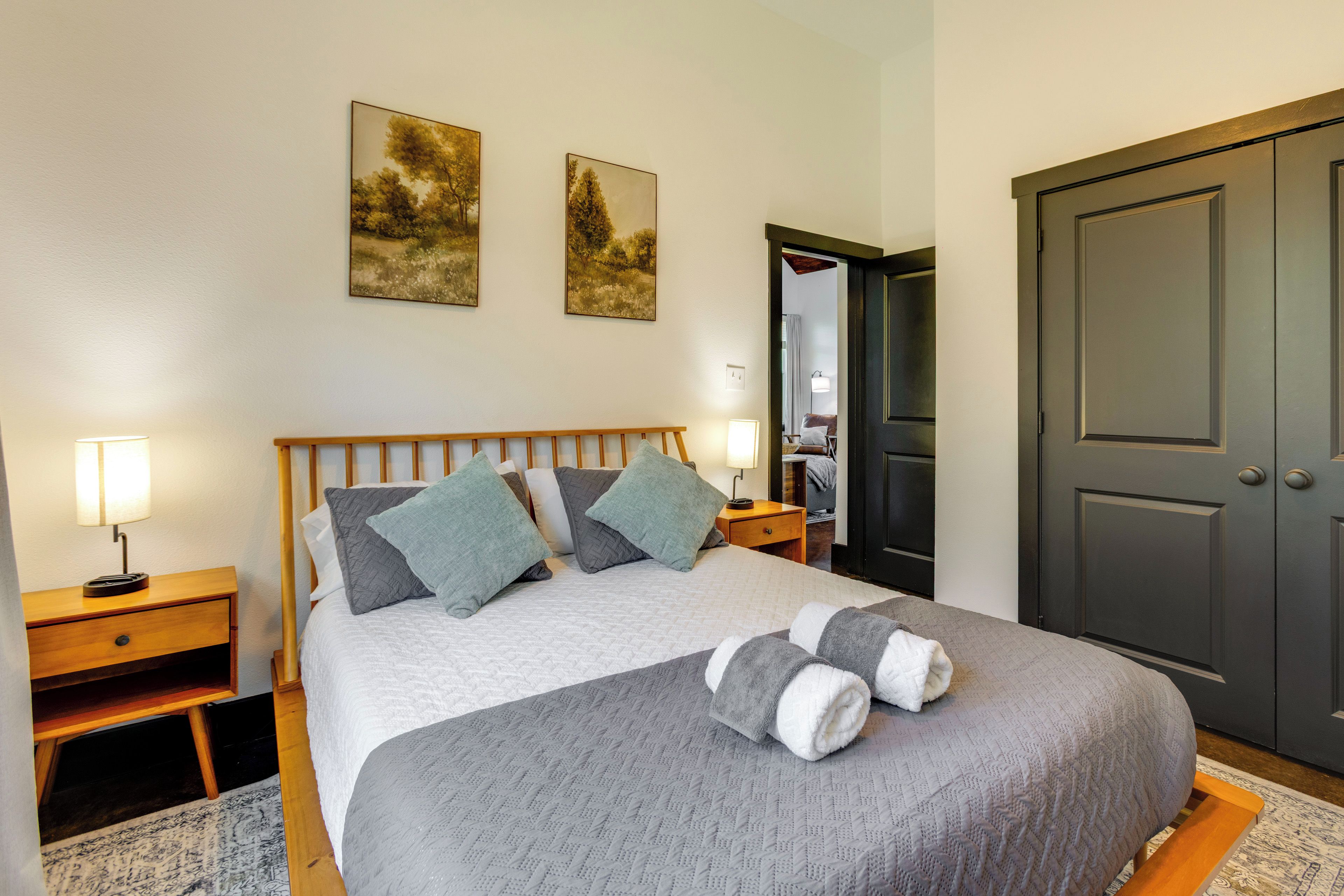 A neatly made bed with a grey blanket and pillows, a bedside table with a lamp, and two paintings on the wall.
