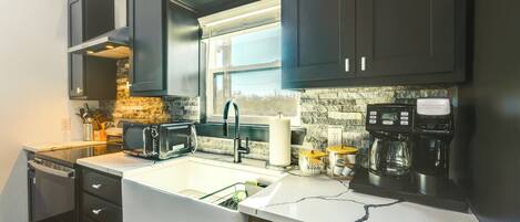 A modern kitchen with dark cabinets, a white countertop, a built-in oven, a microwave, a coffee maker, and a window above the sink.