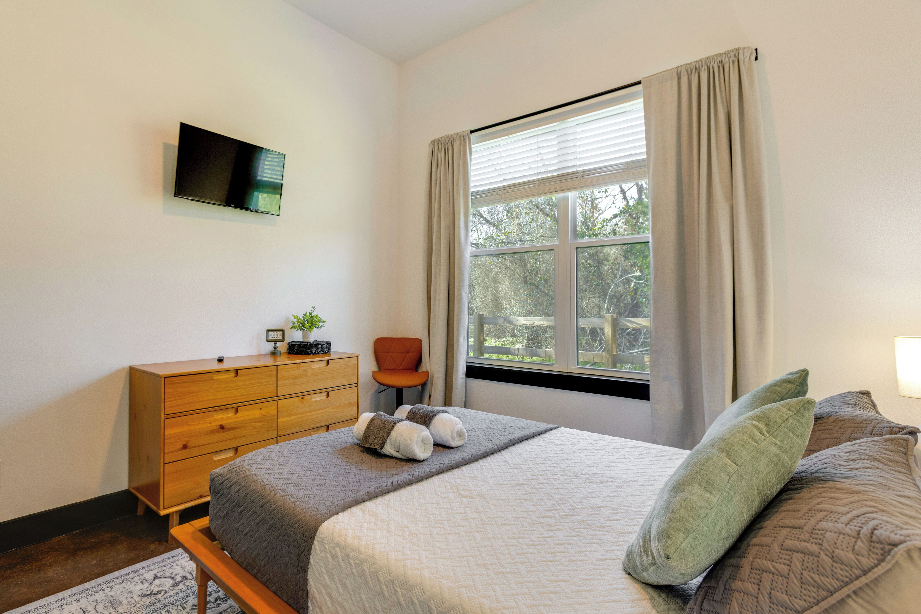 A bedroom with a bed, a wooden dresser, a television on the wall, a window with curtains, and a chair.