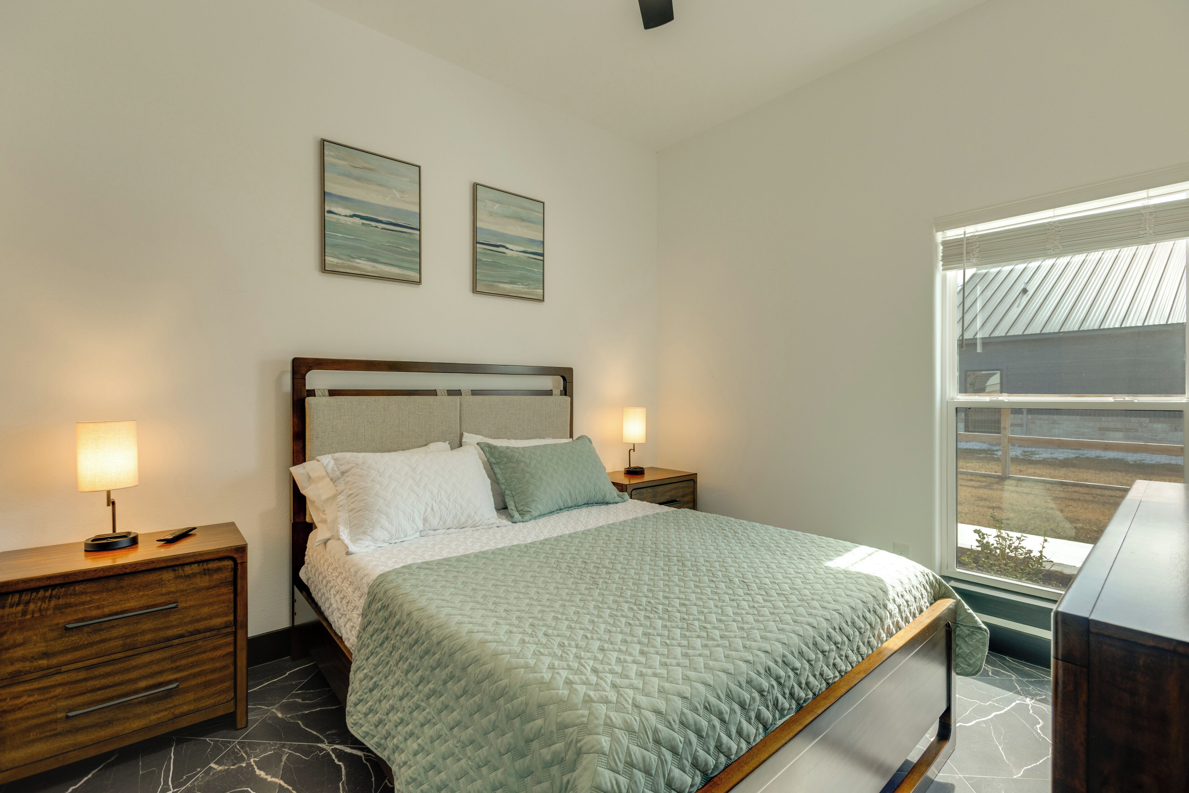 A bedroom with a bed, two bedside tables, a nightstand, a window with blinds, and two wall-mounted paintings.