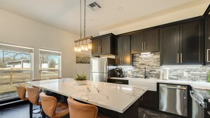 A modern kitchen with dark cabinets, a large island with bar stools, stainless steel appliances, and a view through large windows. - Lake Somerville Family Retreat: 'the Lazy Joey'! (Somerville)