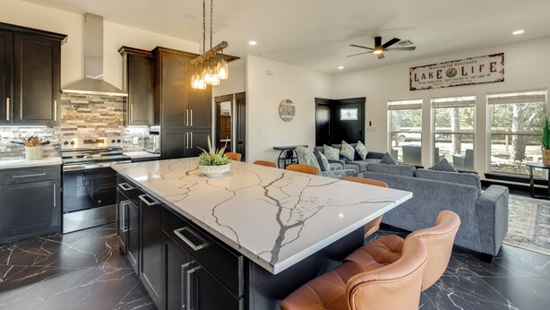 A modern kitchen with dark cabinets, a marble island, and a stone backsplash. There is a dining area with a gray sofa and a ceiling fan. - Lake Somerville Family Retreat: 'the Lazy Joey'! (Somerville)