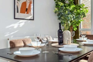 Une table à manger dressée avec de l’assiette blanche, des bols et des verres à vin, une bouteille de vin et une plante en pot.