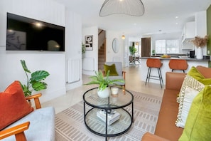 Un salon moderne avec une table basse ronde en verre, un téléviseur à écran plat fixé au mur et un escalier menant à un niveau supérieur.