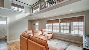 A spacious living room with a sectional sofa, large windows with blinds, a high ceiling, and a visible staircase to an upper level.