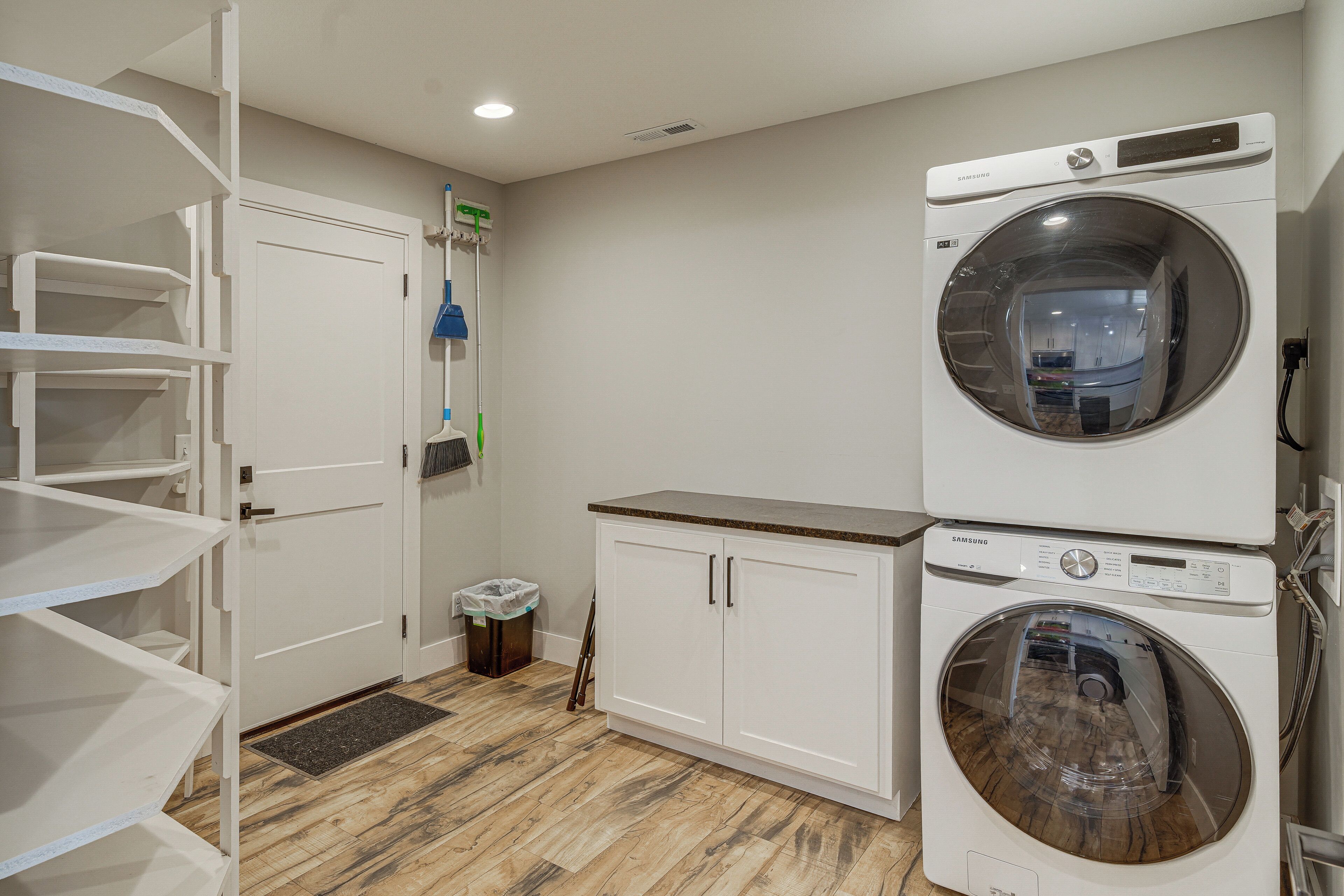 A laundry room with a stacked washer and dryer, a broom, and a trash can.