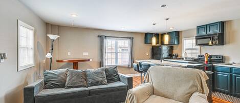 Una sala moderna con un sofá gris, un sillón color beige y un comedor con mesa y sillas. La cocina tiene armarios azul oscuro y un microondas negro.