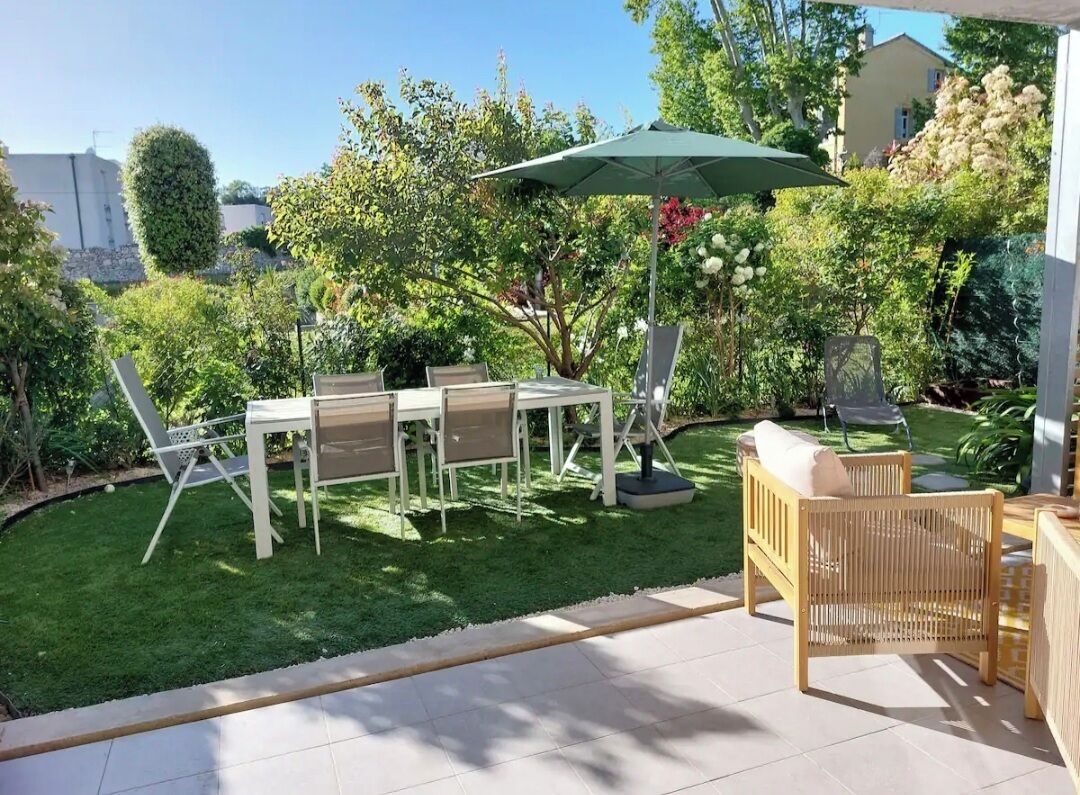 Un espace patio aménagé avec une table et des chaises, un parasol, et un jardin où l’on trouve différentes plantes et arbres.