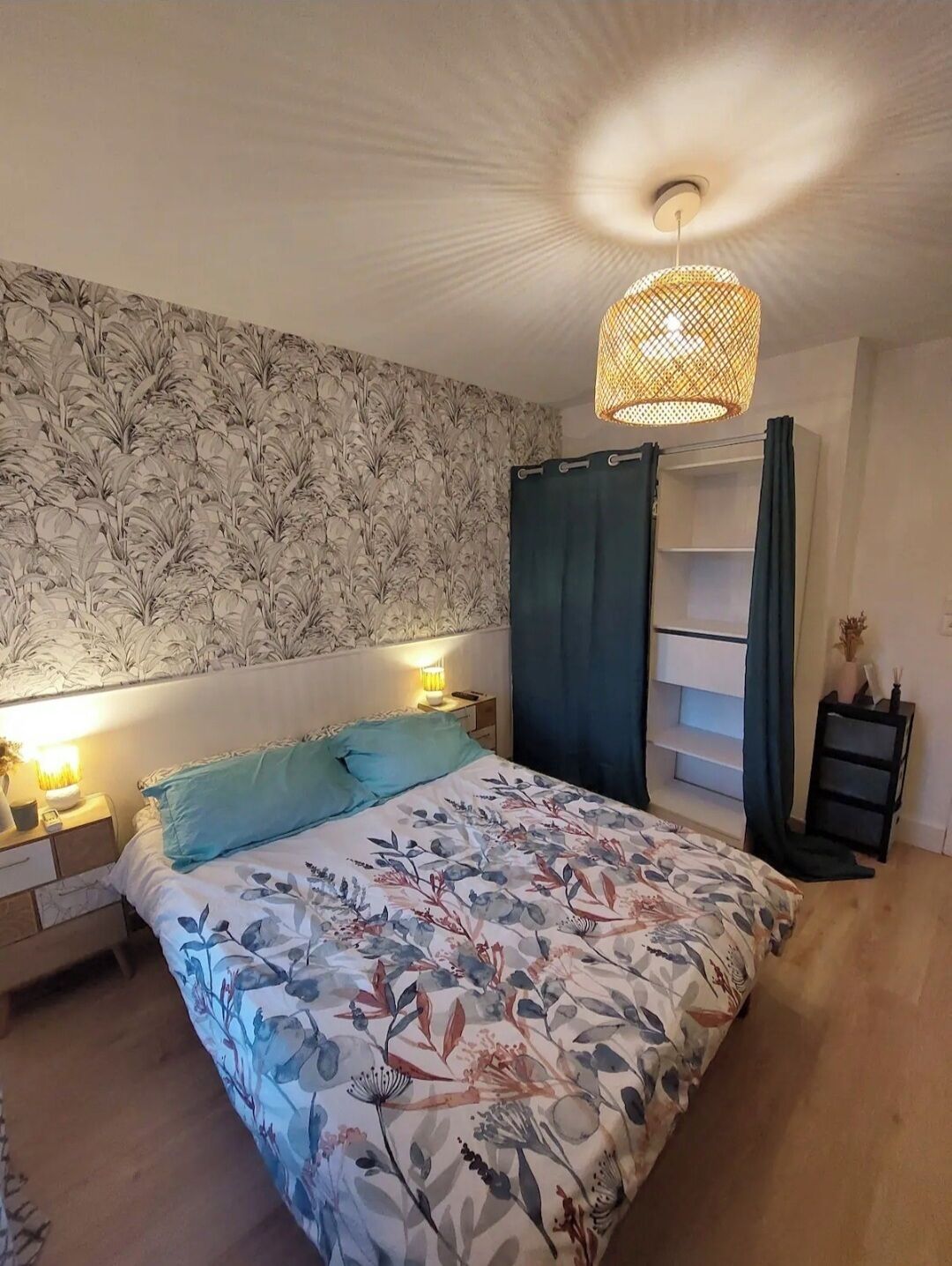 Une chambre à coucher avec une couvre-lit à motifs floraux, un luminaire suspendu, une table de chevet et une étagère.