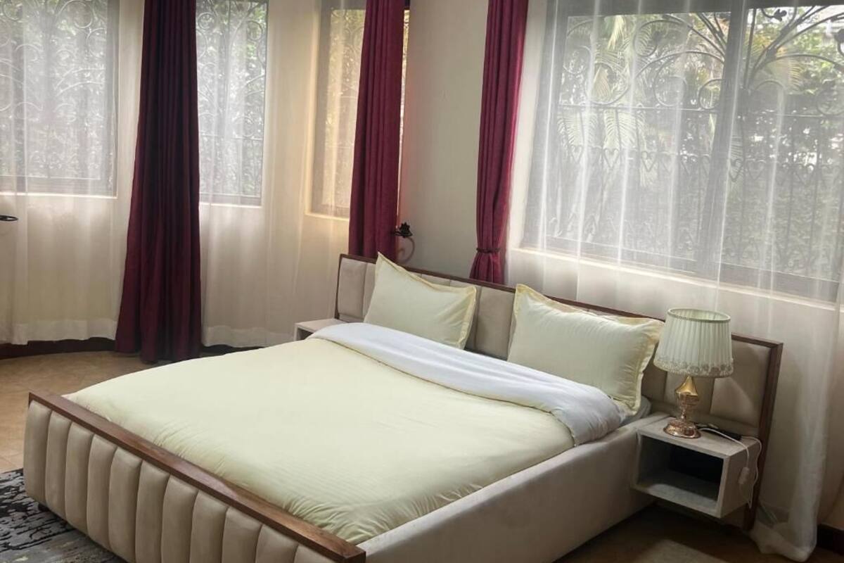 A neatly made bed with a light-colored duvet and pillows, a bedside table with a lamp, and a window with sheer curtains and a view of greenery outside.