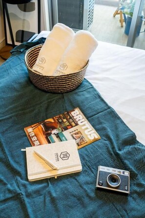 Un lit recouvert d’une courtepointe turquoise, un panier rempli de serviettes pliées, un magazine, un carnet et un appareil photo.