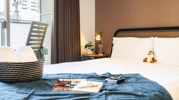 A hotel room with a bed, a nightstand with a lamp, a window with a view of buildings, a chair, and a basket of towels. - Chuan House Kuromon (Osaka)