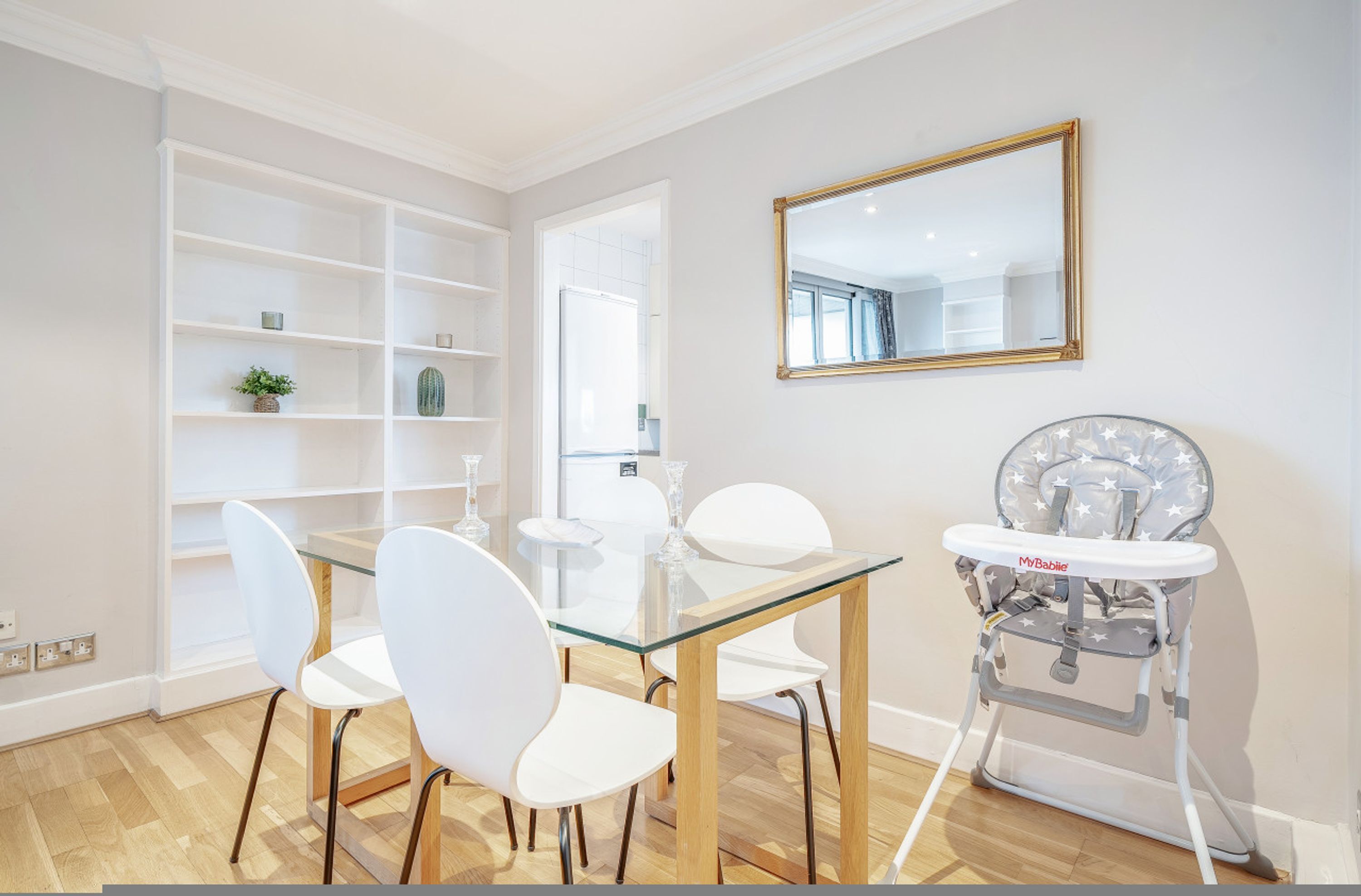 A modern dining area with a glass-top table, white chairs, a high chair, and built-in shelving.