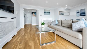 A modern living room with a glass coffee table, a beige sofa with blue and white cushions, wooden flooring, and a flat-screen TV mounted on the wall. - Stunning Flat in Chelsea Kings Road With Balcony (London)