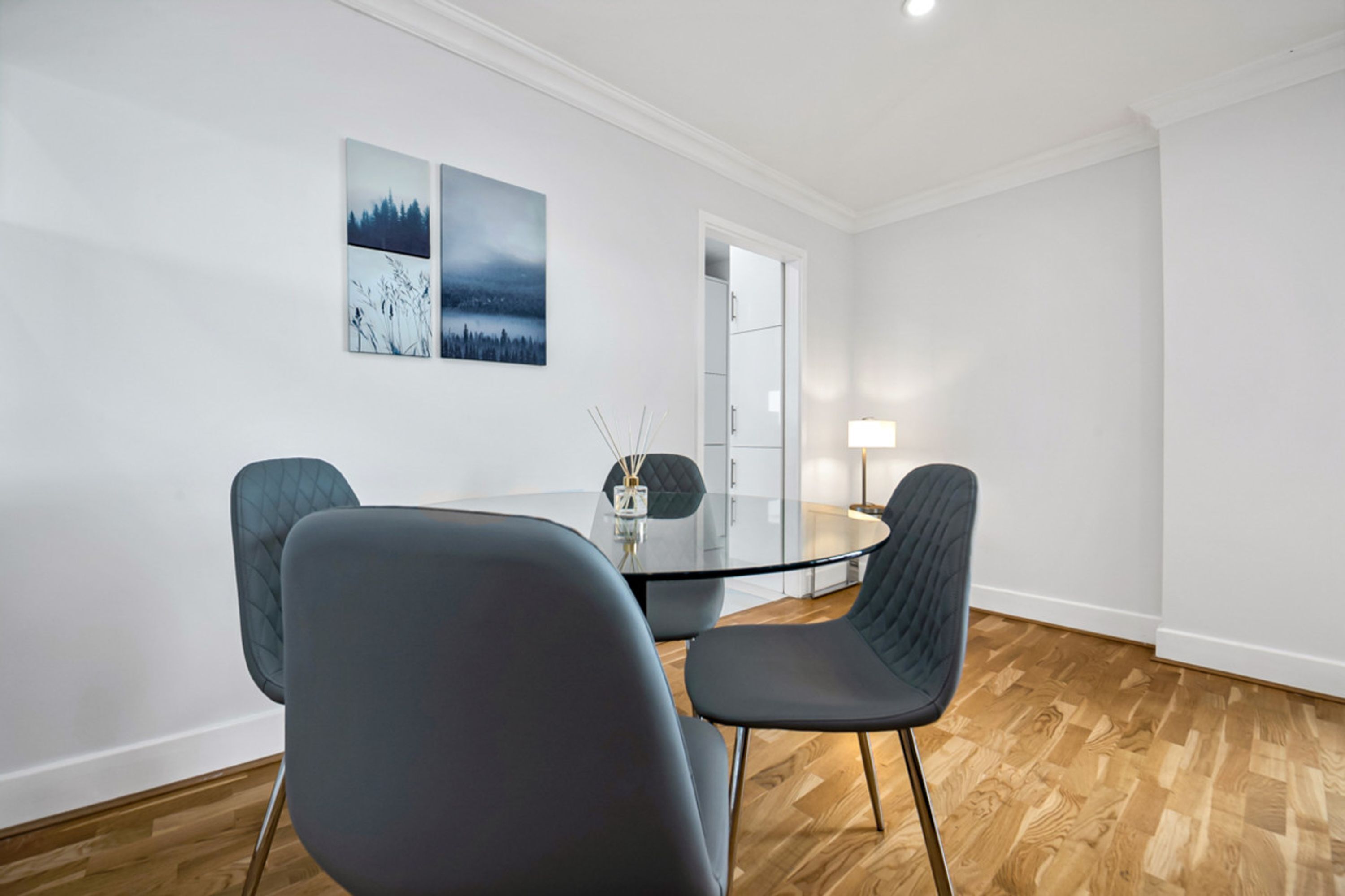 Un comedor moderno con una mesa de vidrio y cuatro sillas, una pared blanca con tres cuadros enmarcados y suelo de madera.