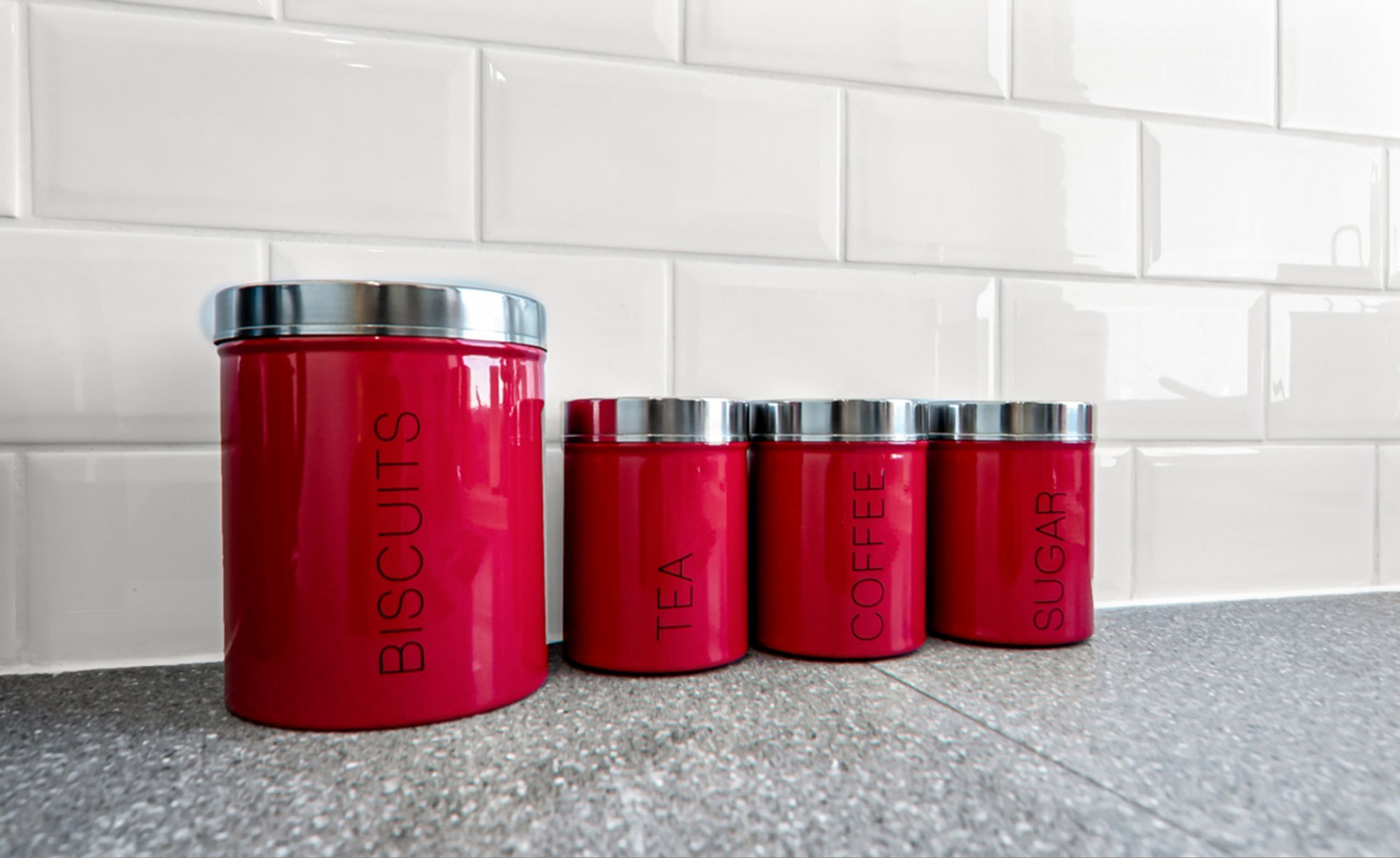 Cuatro recipientes rojos con tapas plateadas etiquetados como “BISCUITS”, “TÉ”, “CAFÉ” y “AZÚCAR” sobre un piso de baldosas, frente a una pared de baldosas blancas.