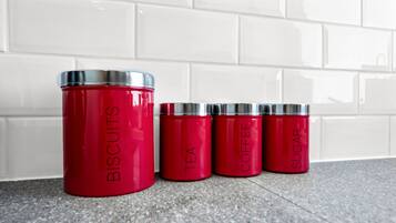 Cuatro recipientes rojos con tapas plateadas etiquetados como “BISCUITS”, “TÉ”, “CAFÉ” y “AZÚCAR” sobre un piso de baldosas, frente a una pared de baldosas blancas.