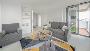 A modern living room with a grey sofa, a glass coffee table, and a patterned rug. There is a white cabinet with shelves, a door leading to another room, and a glass door to an outdoor area. - Stunning Flat in Chelsea Kings Road With Balcony (London)