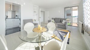 A modern living room with a glass dining table, grey sofa, and a kitchen area with white cabinets and a stainless steel range hood. - Stunning Flat in Chelsea Kings Road With Balcony (London)