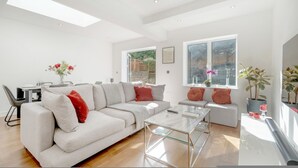 A modern living room with a sectional sofa, a glass coffee table, and a dining area with a table and chairs. There are windows providing natural light and a view of greenery outside. - Maida Vale Gem by Little Venice (London)