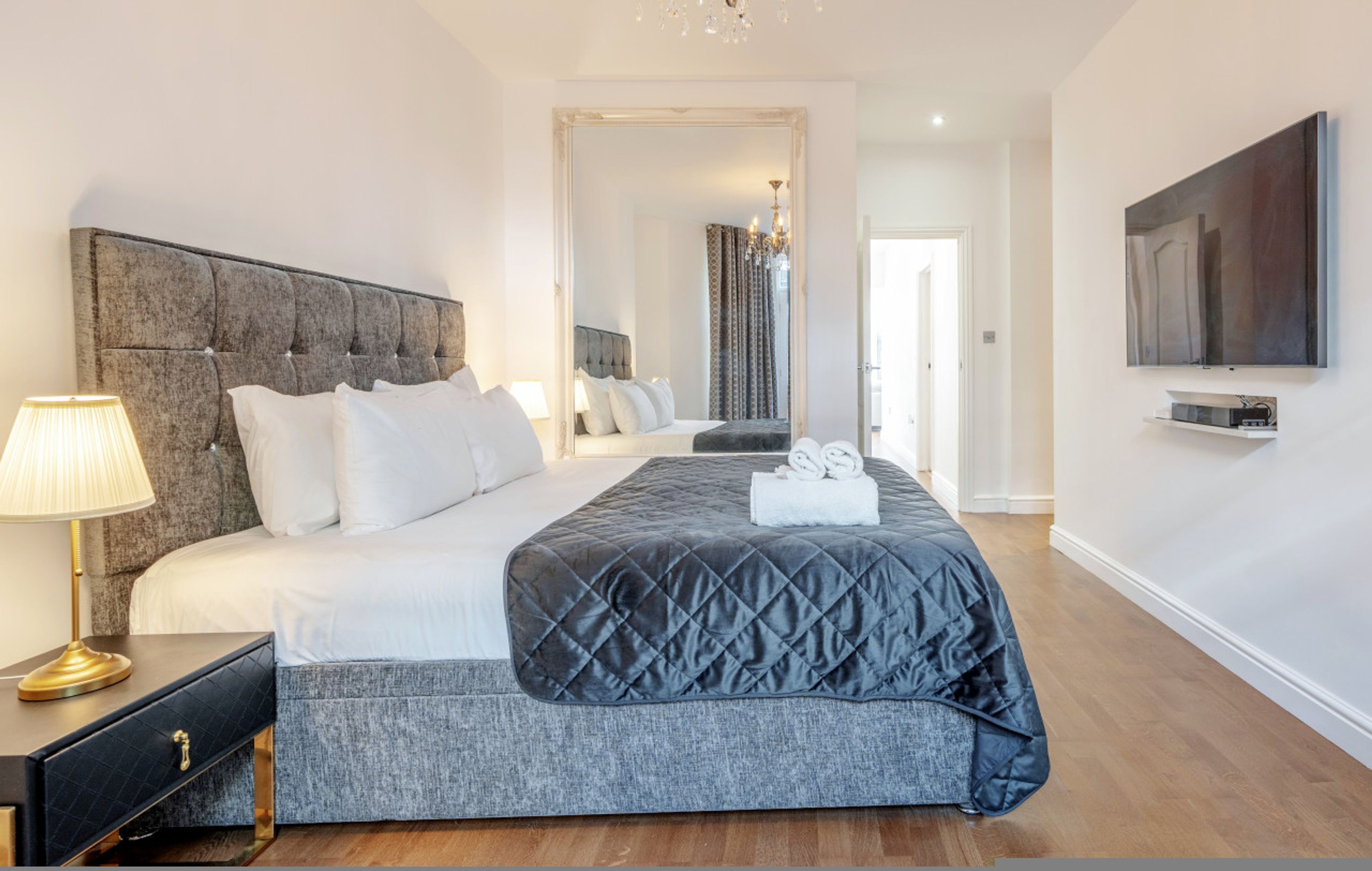 A modern hotel room with a large bed, a bedside table with a lamp, a flat-screen TV mounted on the wall, and a mirror reflecting the room.