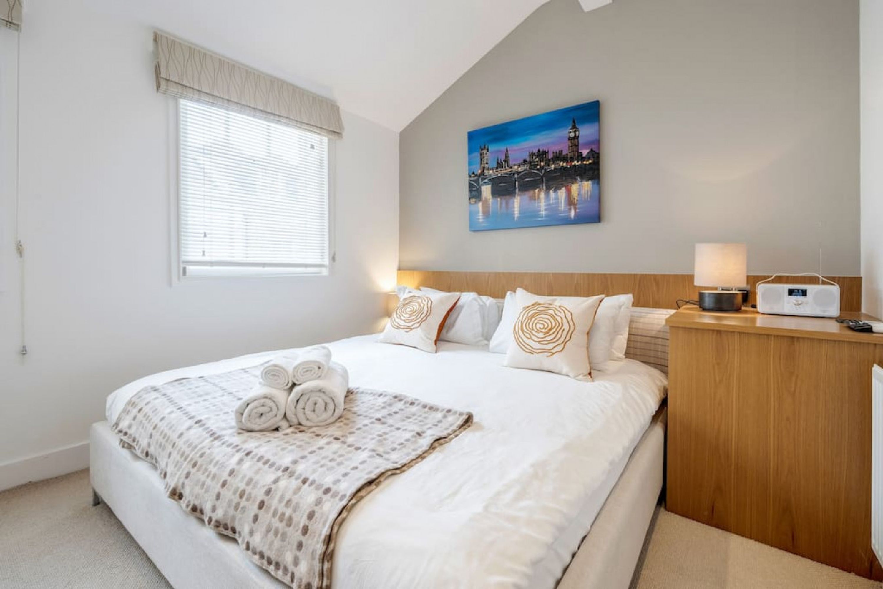 A hotel room with a large bed, a painting of a cityscape, a bedside table with a lamp, and a wooden nightstand.