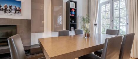 A modern dining room with a large wooden table, six chairs, a fireplace, a bookshelf, and a large window with curtains.