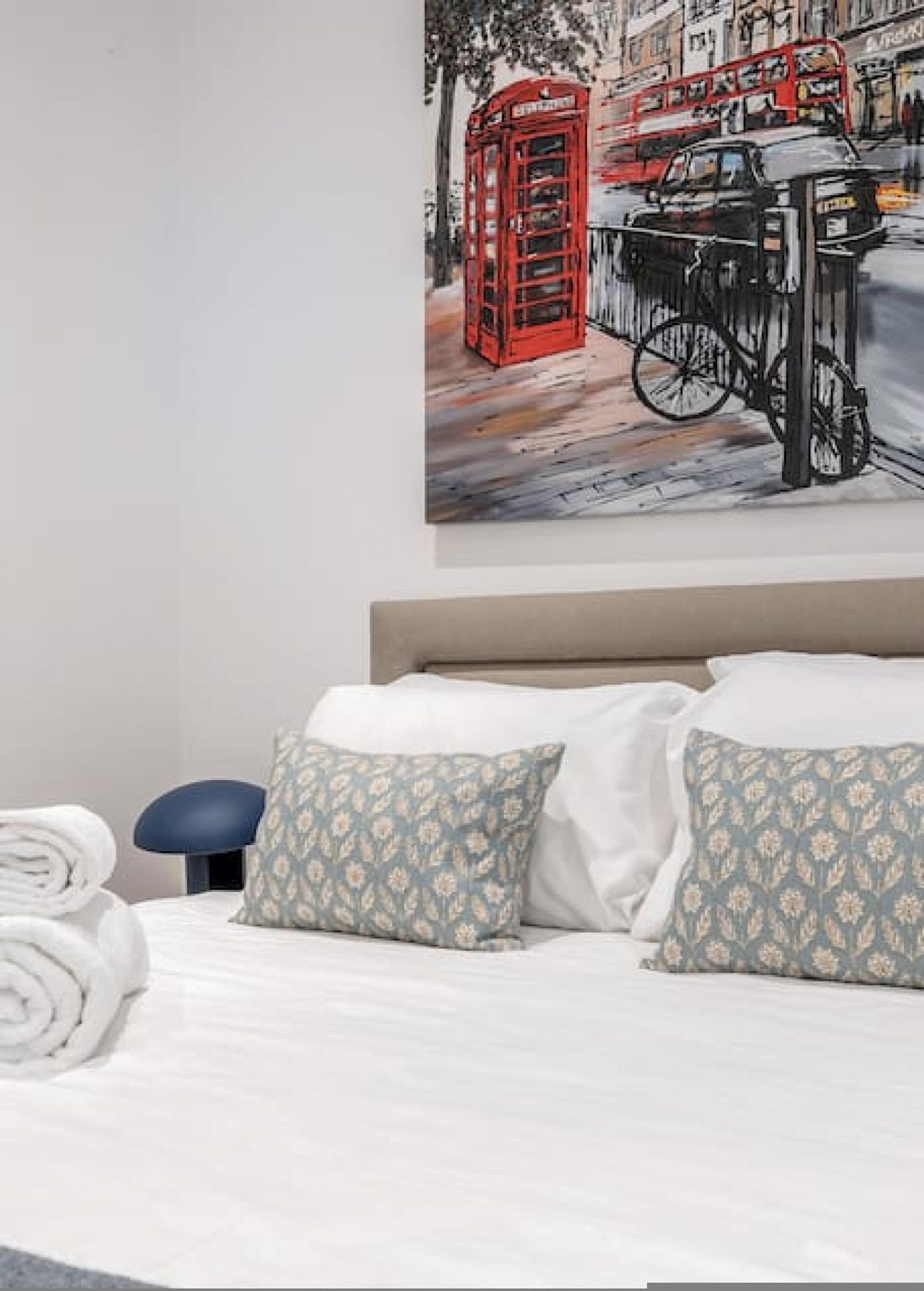 Une chambre d’hôtel avec un lit, des oreillers, une serviette pliée et une œuvre d’art accrochée au mur représentant une cabine téléphonique rouge et une voiture noire.