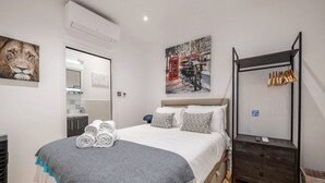 A neatly made bed with rolled towels, a bedside table, a wardrobe, and a wall with a painting of a cityscape. - Conduit Mews Luxurious Mews House Next to Hyde Park (London)