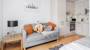 Una sala de estar moderna con un sofá gris, almohadones coloridos, una mesita con una lámpara, y una cocina con armarios y electrodomésticos blancos.