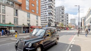 Un taxi negro con una señal amarilla en el techo, circulando por una calle urbana con edificios altos y peatones.
