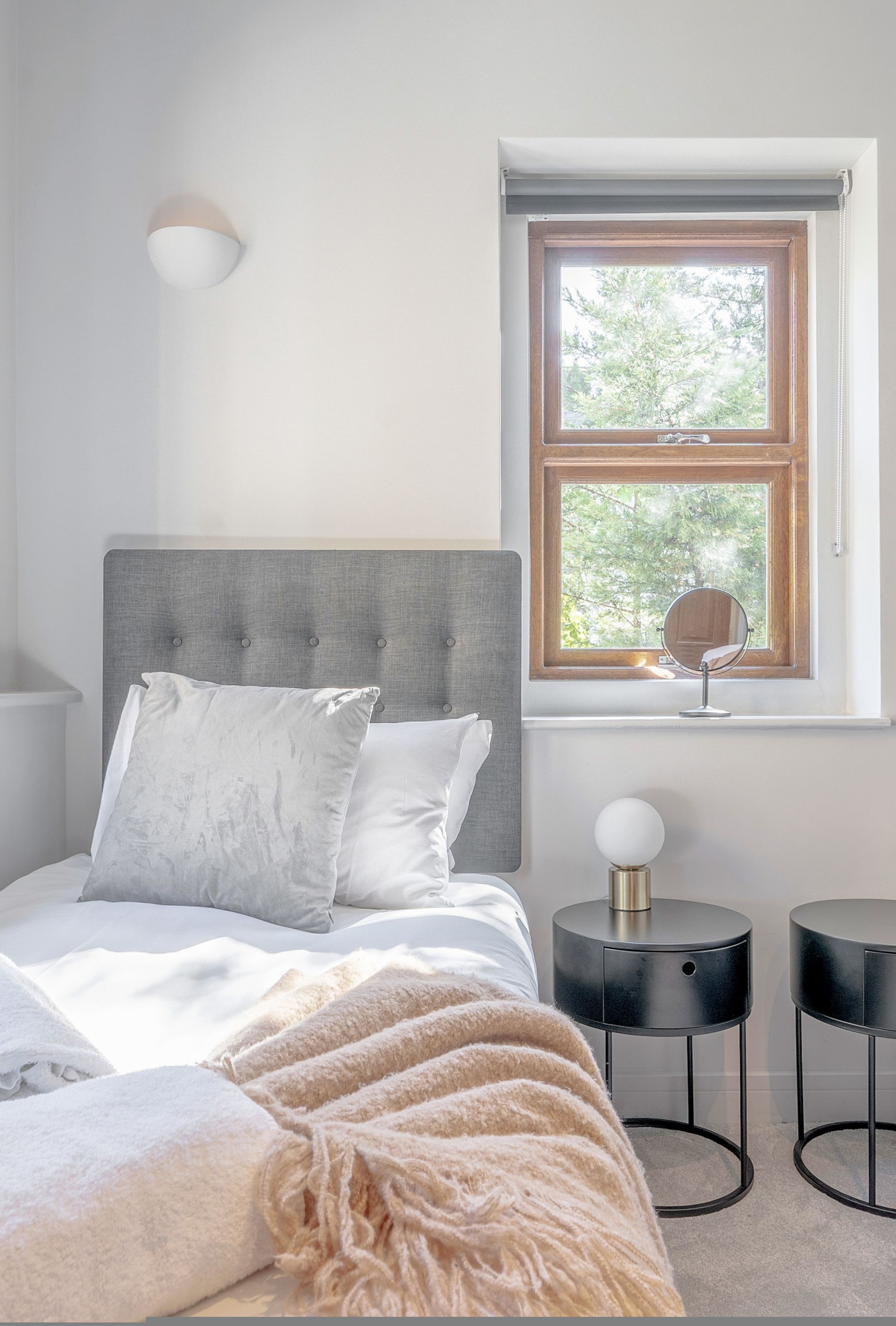 A bedroom with a bed, a bedside table with a lamp, a window with a view, and a wall-mounted light fixture.