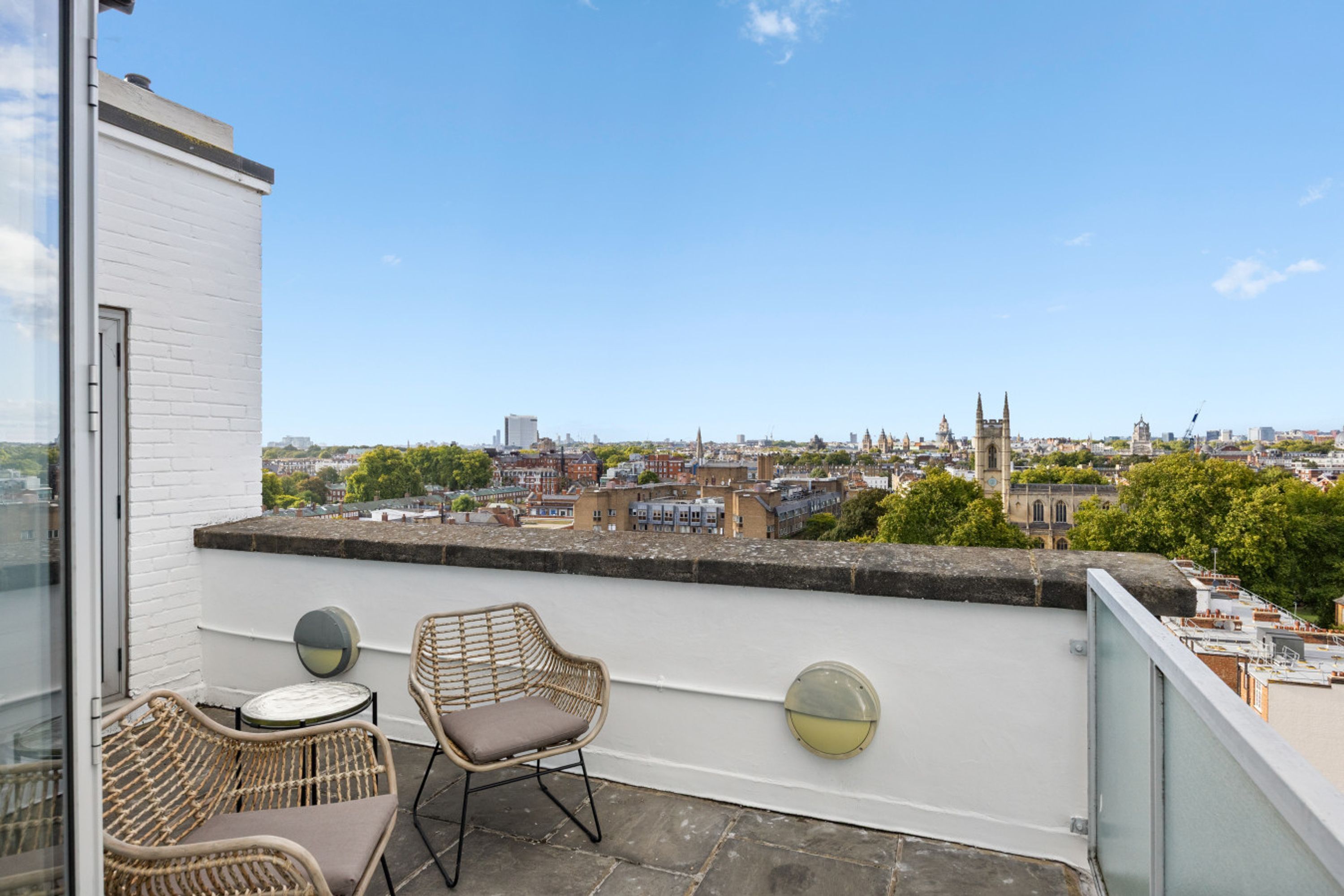 En balkon med fletstole og et bord, med udsigt over en by med historiske bygninger og en kirke.