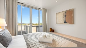 Ein Hotelzimmer mit einem großen Bett, Blick auf die Stadt durch raumhohe Fenster, einem Bild an der Wand und einem Nachttisch mit Lampe und einer Flasche.