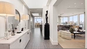 Ein modernes Interieur mit einer langen Diele, die in einen Wohnbereich führt, mit einer zentralen, schwarzen Skulptur, einem großen Fenster und Blick auf die Stadt.