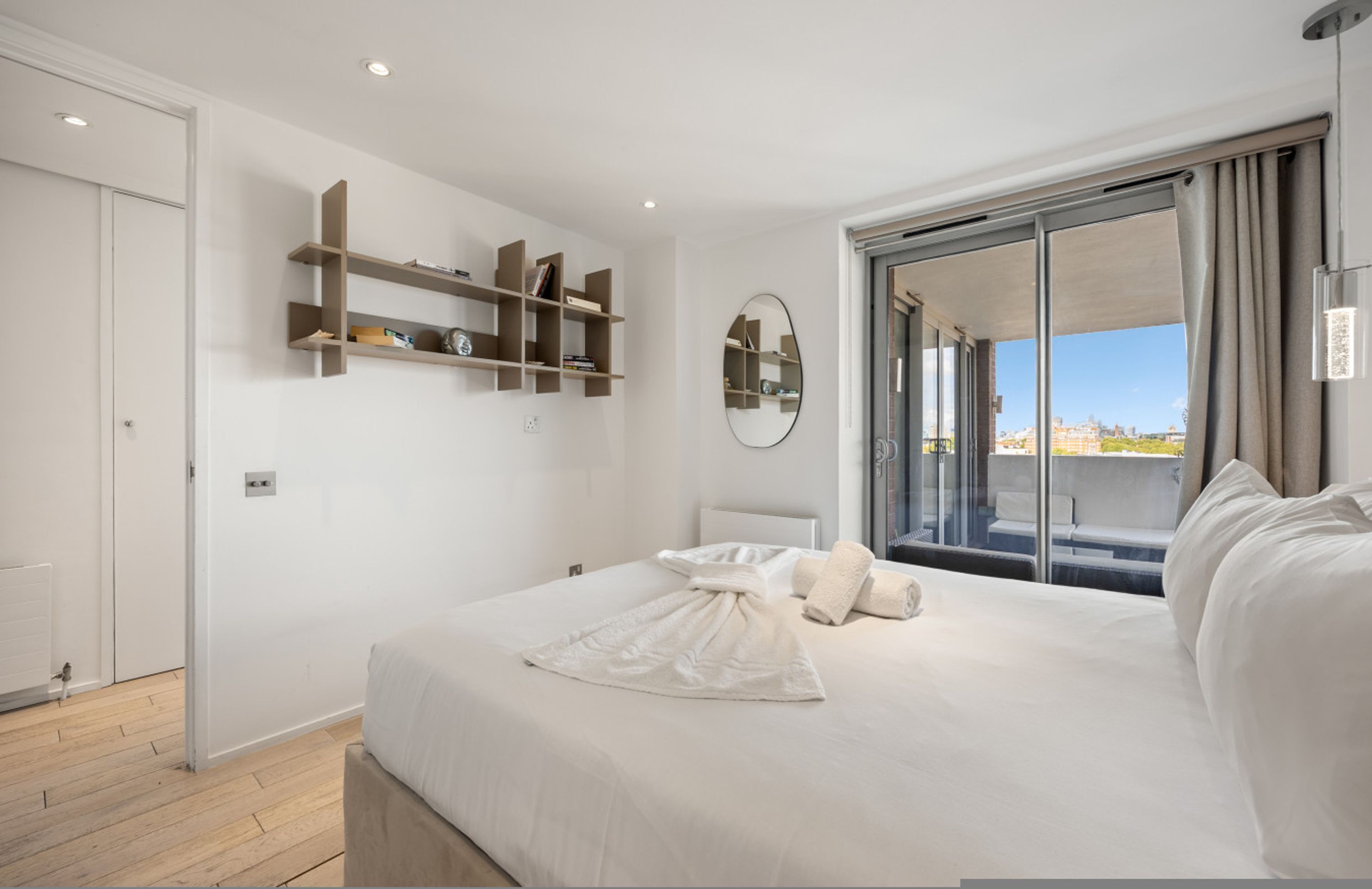 Een moderne hotelkamer met een groot bed, een schuifglasdeur naar het balkon, en ingebouwde planken met boeken en decoratieve objecten.