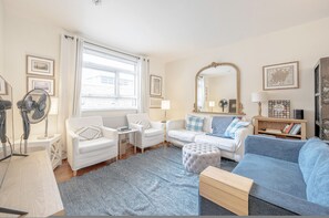 A well-lit living room with a large window, white furniture, a blue sofa, and a mirror. - Cosy Flat in Chelsea (London)