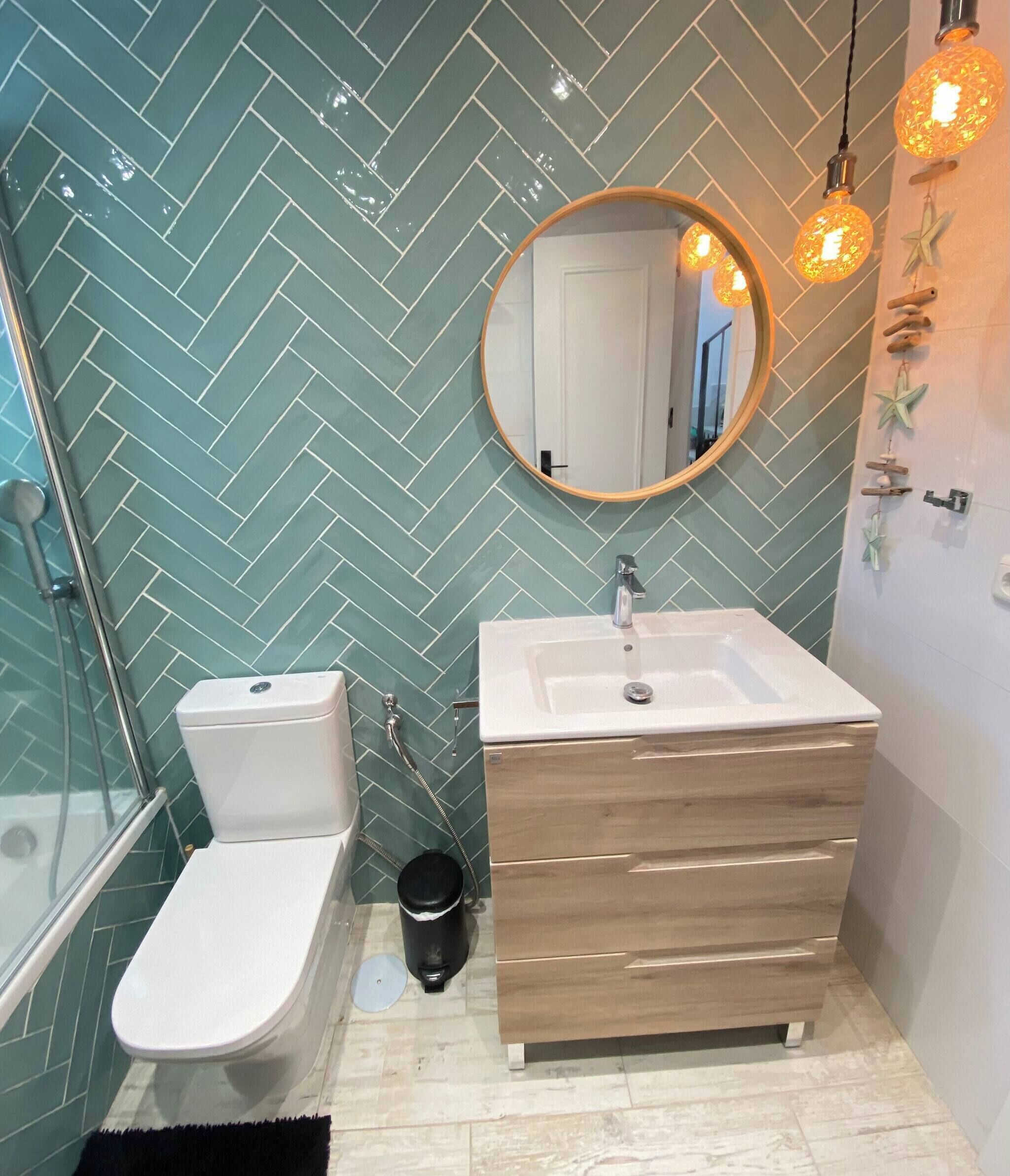 Un baño con salpicadero de azulejos tipo espiga, un espejo redondo, un lavabo blanco y un mueble de tocador de madera.