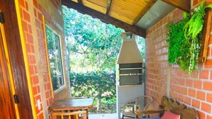 A covered patio with a brick wall, a wooden table, a blue sofa with a red pillow, and a grill. - Tarumã Challes 04, Complete chalet with balcony and barbecue, pets allowed. (Bonito)