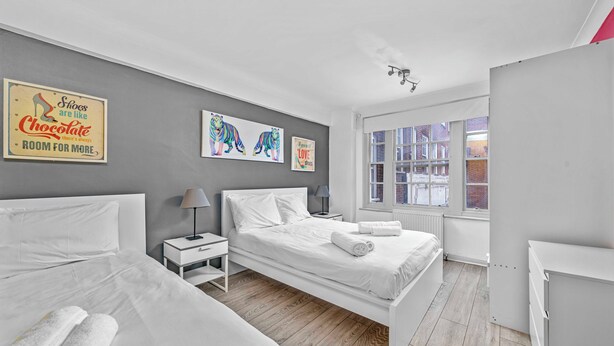 A modern bedroom with a large bed, a smaller bed, a nightstand, a lamp, and a window with a view of buildings. - 1 Bed Flat in London (London)
