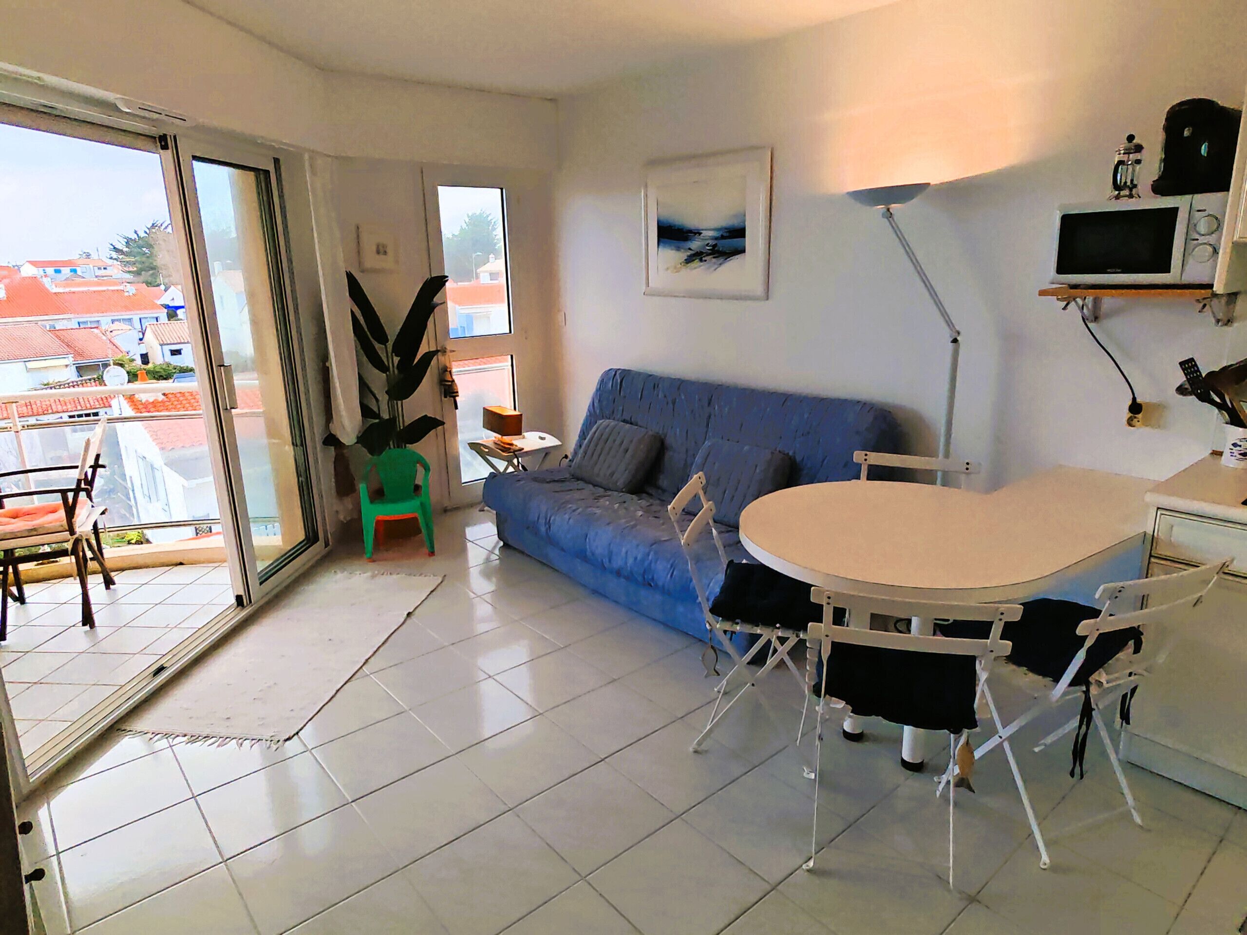 A living room with a blue sofa, a dining table with chairs, a green chair, a large potted plant, and a painting on the wall.