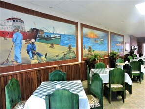 A restaurant with a mural of fishing scenes on the wall, green upholstered chairs, and checkered tablecloths. - Ngandu at Sea  (Walvis Bay)