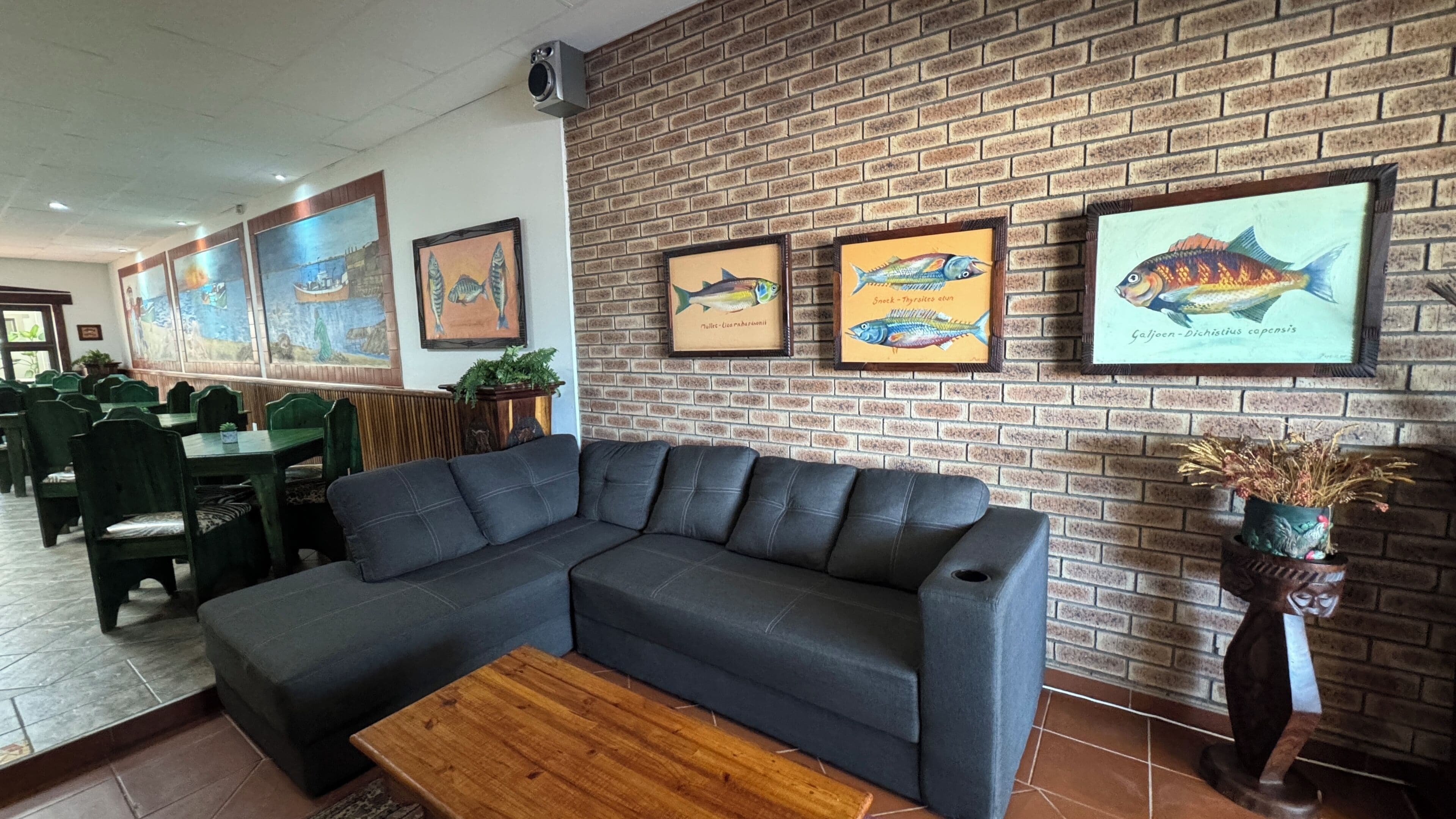 Interior de restaurante com parede de tijolos, quadros de peixes emoldurados, um sofá cinza e uma mesa de centro de madeira.