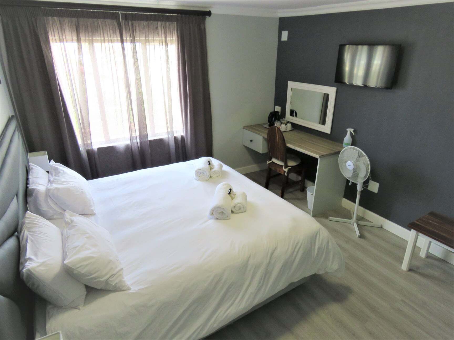 Una habitación de hotel con una cama, un escritorio, una silla, un espejo y un ventilador de pie.
