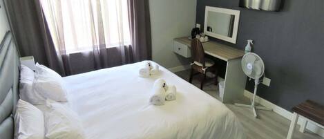 Una habitación de hotel con una cama, un escritorio, una silla, un espejo y un ventilador de pie.